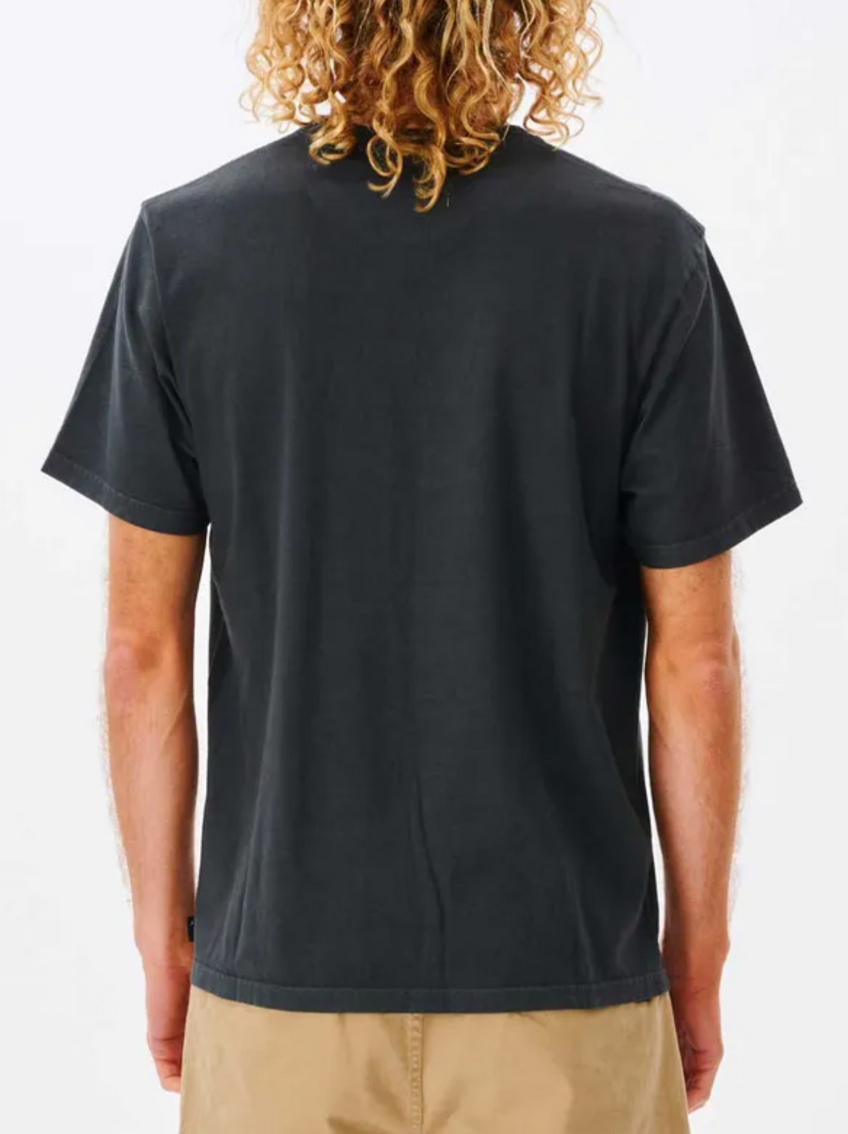 A person with curly, shoulder-length hair is seen from behind, wearing a relaxed fit black Rip Curl Plain Wash Tee and light brown shorts against a white background.