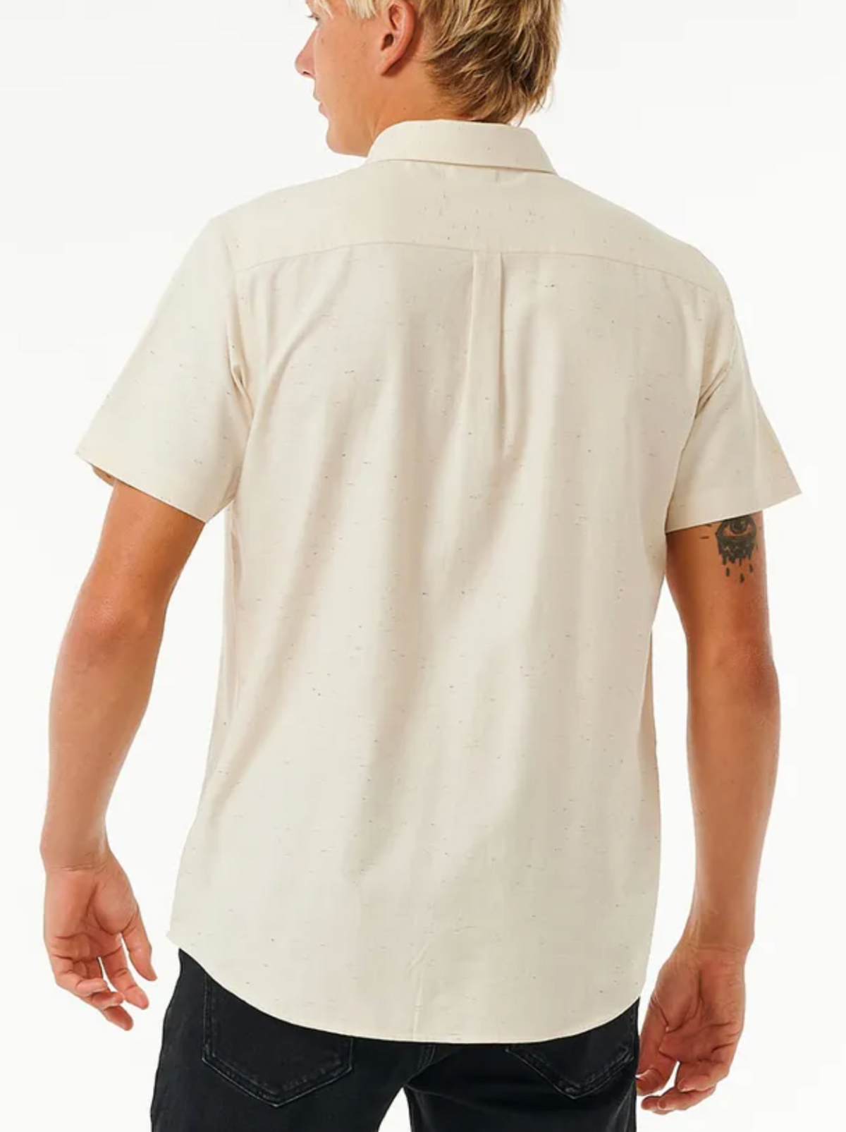 A person with short blond hair is wearing a Rip Curl Ourtime Short Sleeve Shirt in light beige and black pants, facing away from the camera. A tattoo is visible on their left arm.