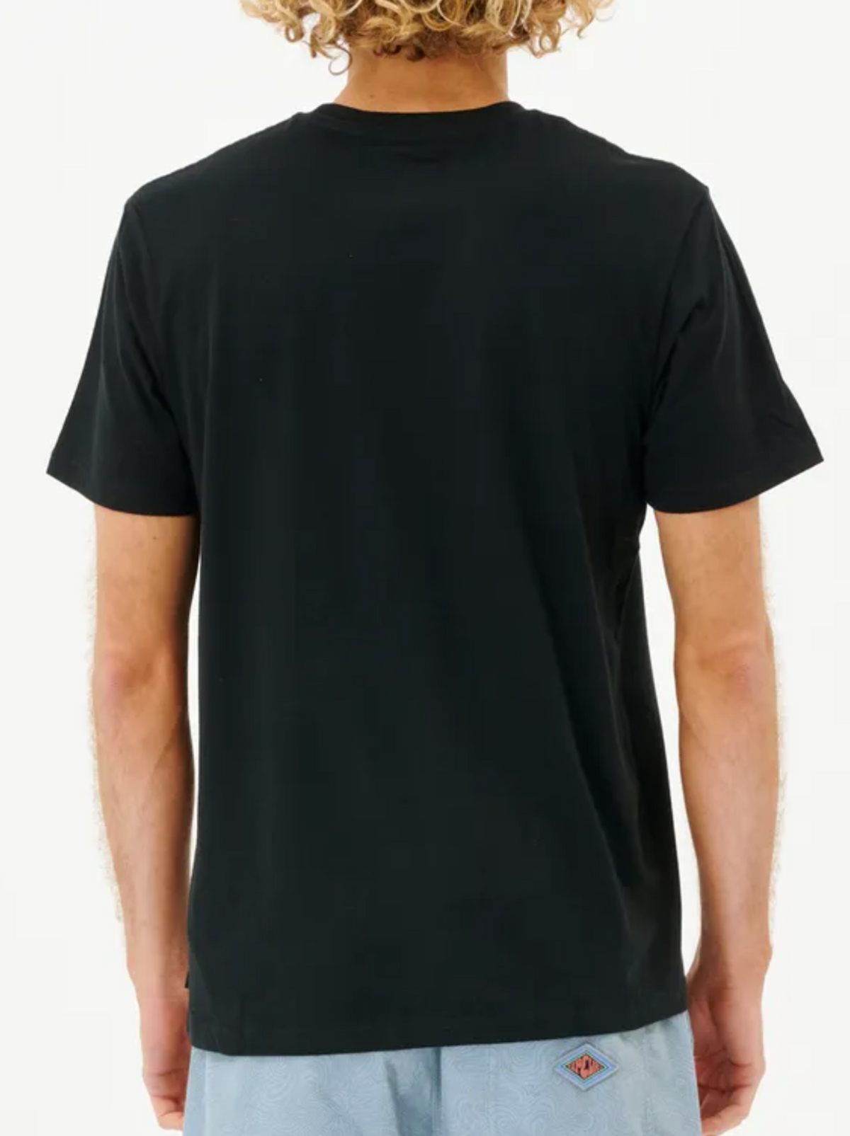A person with curly hair is seen from behind, wearing a classic Rip Curl Plain Pocket Tee made of 100% cotton in black and paired with light blue shorts, set against a plain white background.