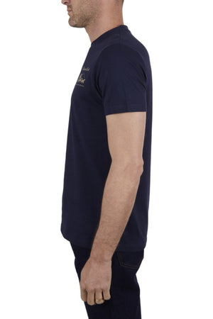 A man stands in profile facing left, arms relaxed by his side, wearing the Thomas Cook Coach Tee in navy blue cotton jersey and dark jeans. The background is plain white.