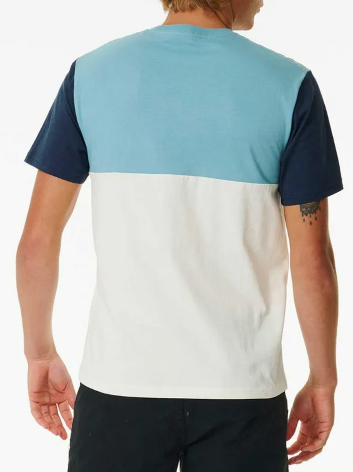 A person wears a Rip Curl Surf Revival Custom Tee, featuring 100% cotton and a color-block design with a blue upper, white lower, and dark navy sleeves. Facing away, their left arm tattoo is visible. With light brown hair and matching black pants, they exude an effortless laid-back vibe.