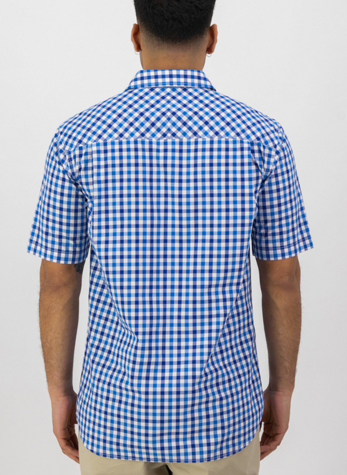 A man stands with his back to the camera, wearing a Swanndri Moreau Short Sleeve Shirt in blue and white check pattern and beige pants. Made from 100% cotton in a tailored fit, he is set against a plain white background.