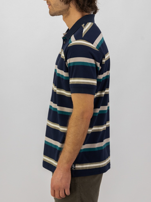 A man with wavy brown hair, dressed in dark pants and the Swanndri Winfred Stripe Polo, stands sideways against a plain light background.
