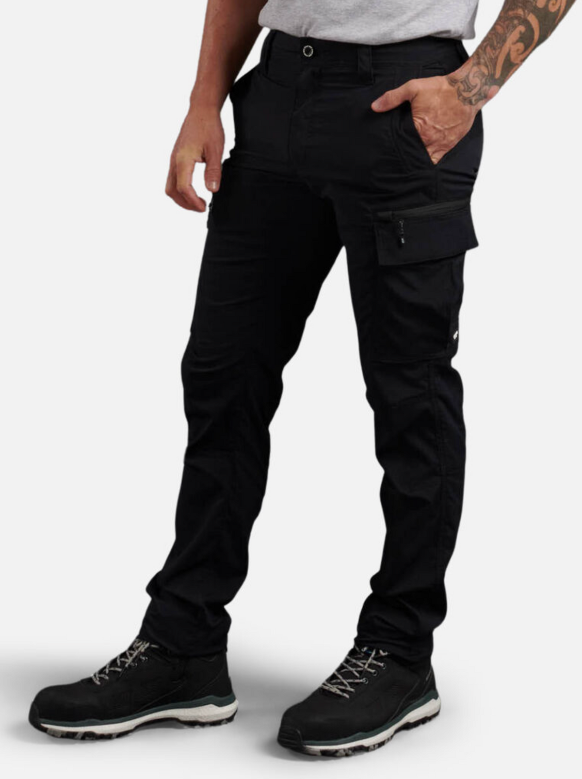 A person wears black King Gee Trademark Cargo Pant featuring moisture control, paired with a grey t-shirt and black hiking shoes. One hand is in the front pocket, the other arm shows a tattoo. The image is cropped at the shoulders and feet.