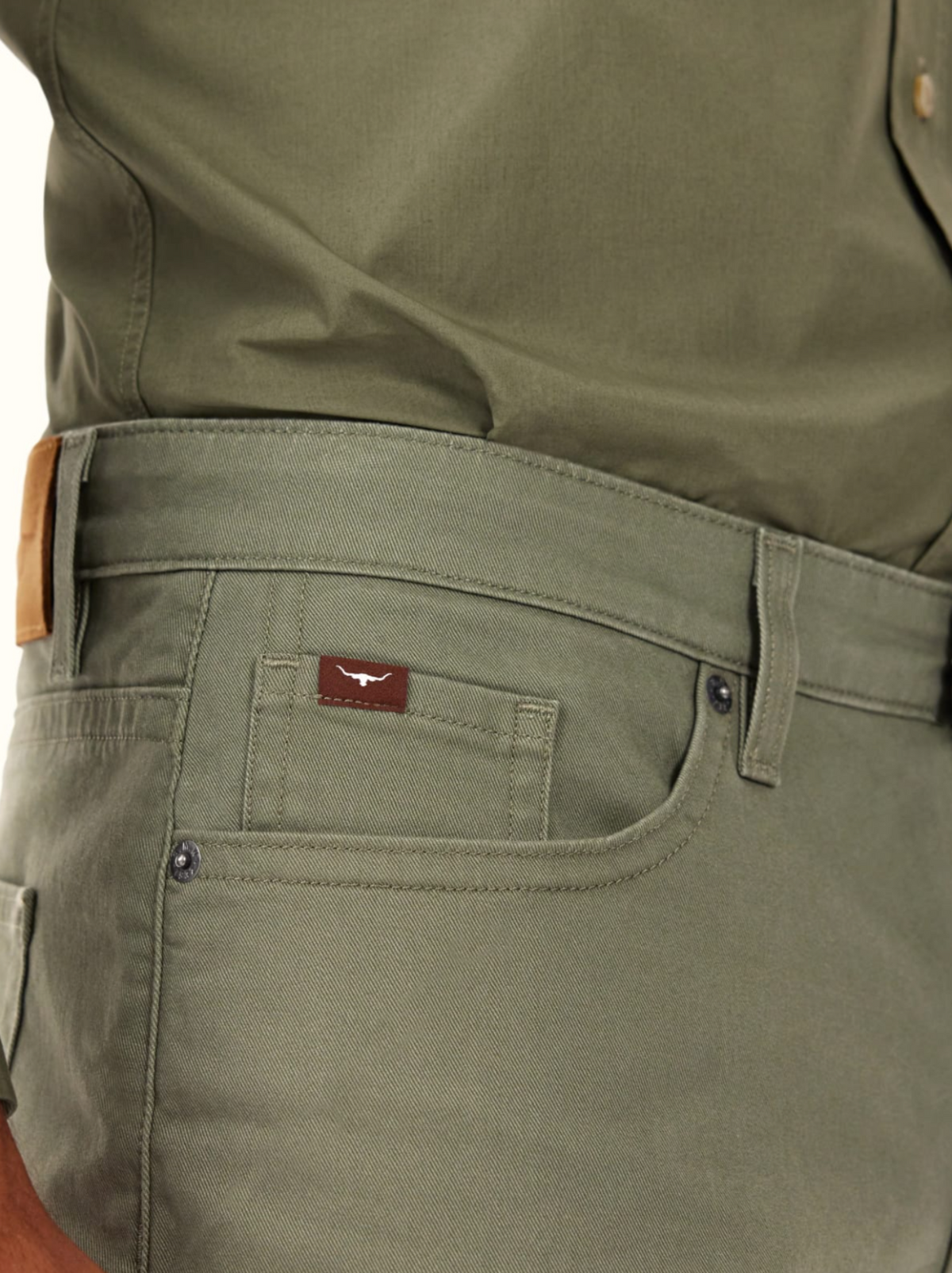 Close-up of someone in RM Williams Nicholson Short—a smart-casual olive green stretch cotton pant with a brown leather patch on the waistband, front pocket, belt loops, and button—paired with a matching green shirt tucked in.