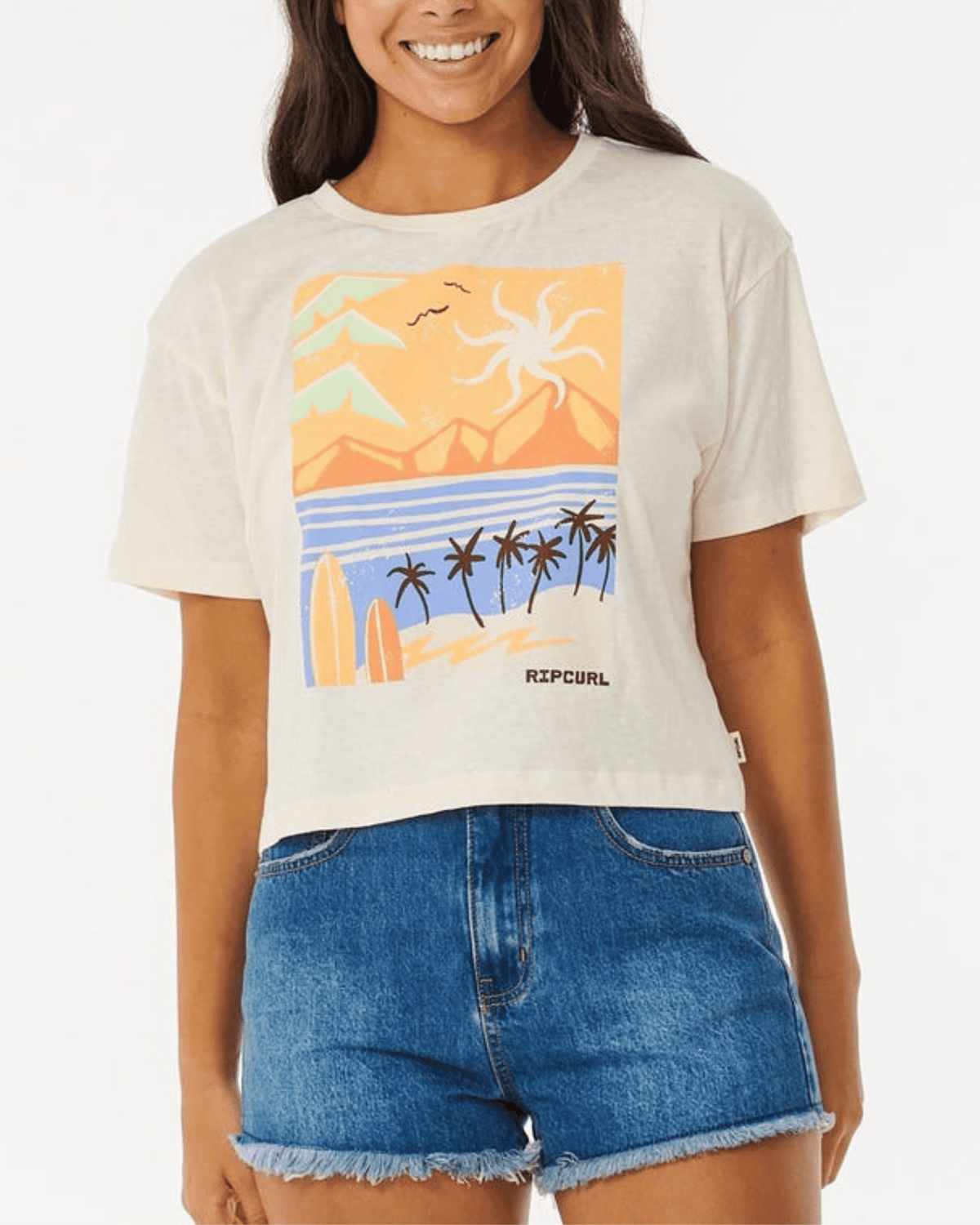 Wearing the Rip Curl Calypso Crop Tee with a vibrant beach and palm tree graphic, a woman smiles and pairs it stylishly with blue denim shorts.