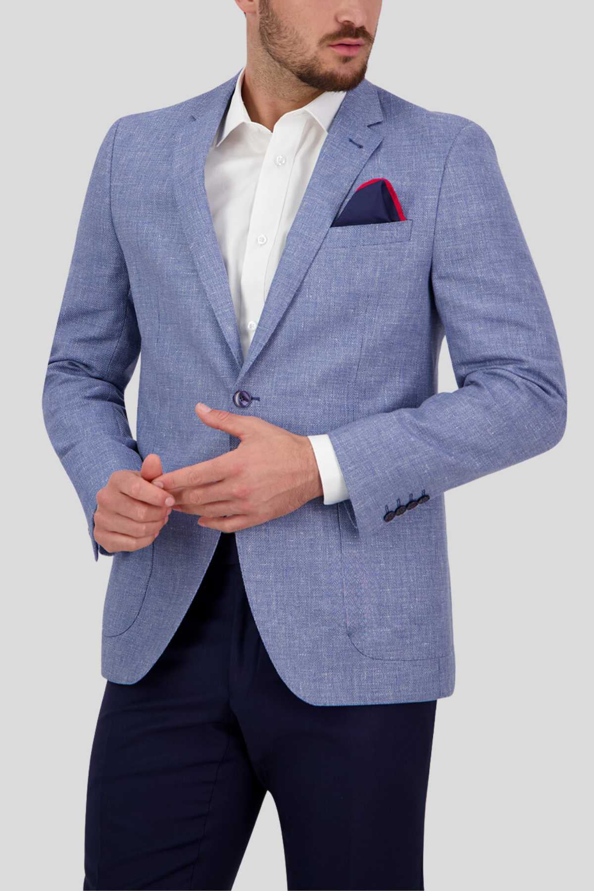 A man is dressed in a Bruton Asher Linen Blazer in light blue, paired with a white dress shirt and dark trousers. A red pocket square adorns the breast pocket of the professional formal blazer. With his hands positioned in front of him, he gazes slightly to the side against a plain gray background.