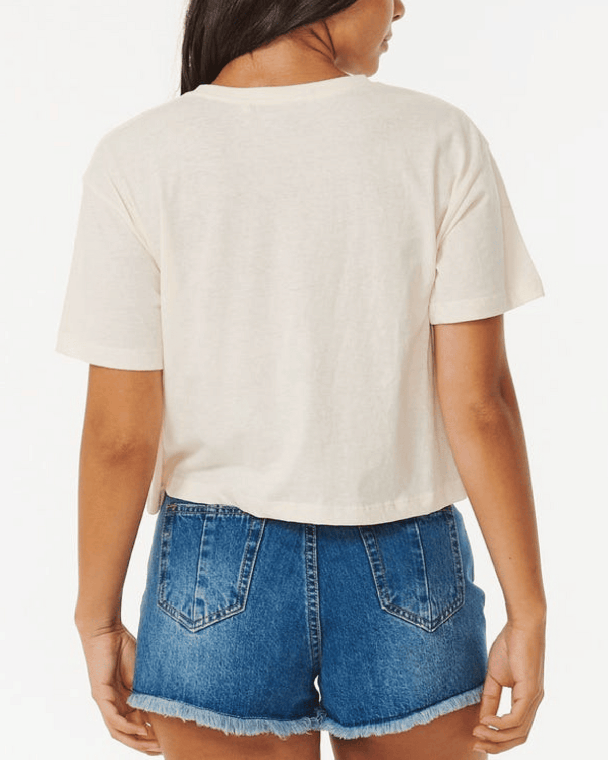 A person is shown from the back wearing the Rip Curl Calypso Crop Tee with light-colored short sleeves and blue frayed-hem denim shorts against a plain white background.