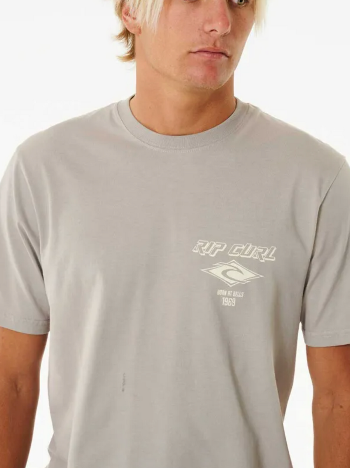 A young man with blonde hair models the Rip Curl Fade Out Icon Tee, a light grey cotton t-shirt with a small white logo and text on the left chest, standing against a plain white background.