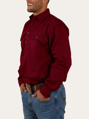 A man wearing a maroon Ringers Western King River Half Button Shirt with chest pockets, blue jeans, and a decorative belt buckle stands with his hands in his pockets and smiles.