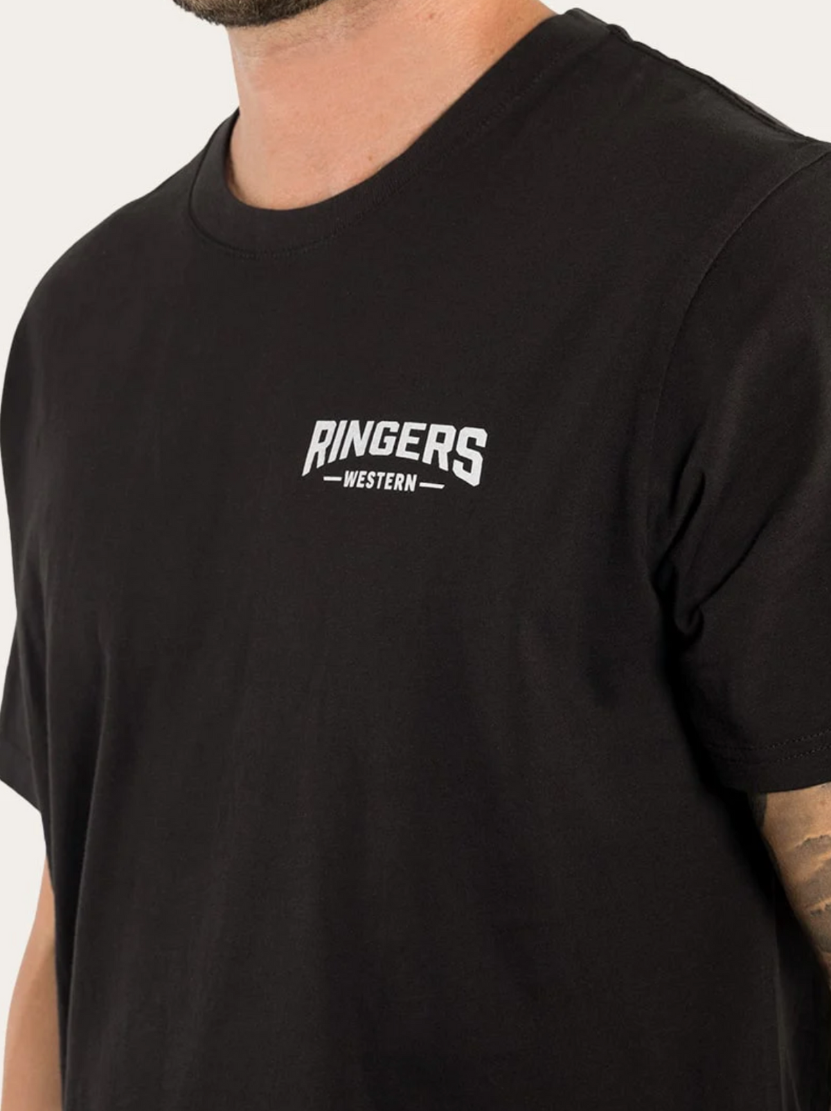 A person wearing a Ringers Western Squadron Loose Fit T-Shirt, characterized by its black color and the white "RINGERS WESTERN" logo on the left side of the chest. The loose fit design, crafted from 100% cotton, ensures comfort. The tattoo on the person's right arm is partially visible against a plain, light-colored background.