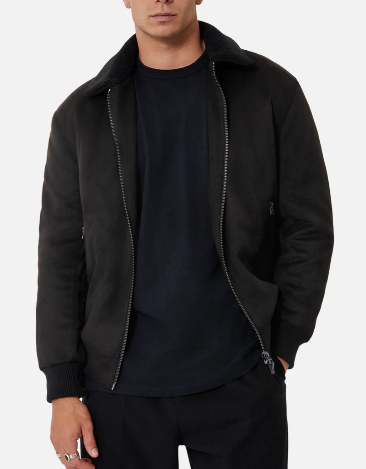 A man wearing the Industrie The Mig Jacket—a black zip-up with a faux sherpa collar, ribbed cuffs, and sleek suede outer—stands with one hand in his pocket against a plain background.