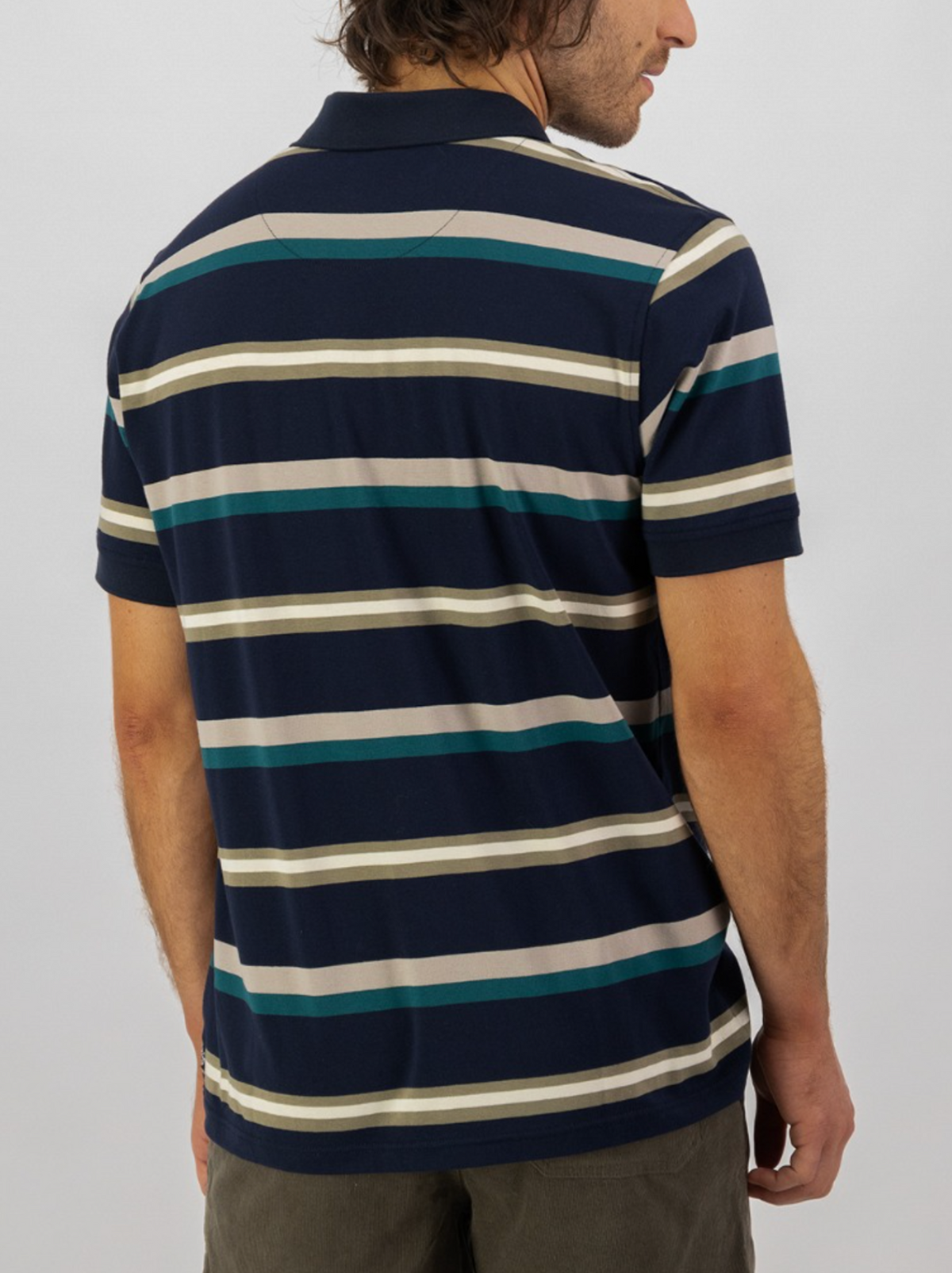A man, shown from the back with his head turned, wears the Swanndri Winfred Stripe Polo—a navy blue polo made from cotton and elastane, featuring white, beige, and teal horizontal stripes for a comfortable yet stylish look.