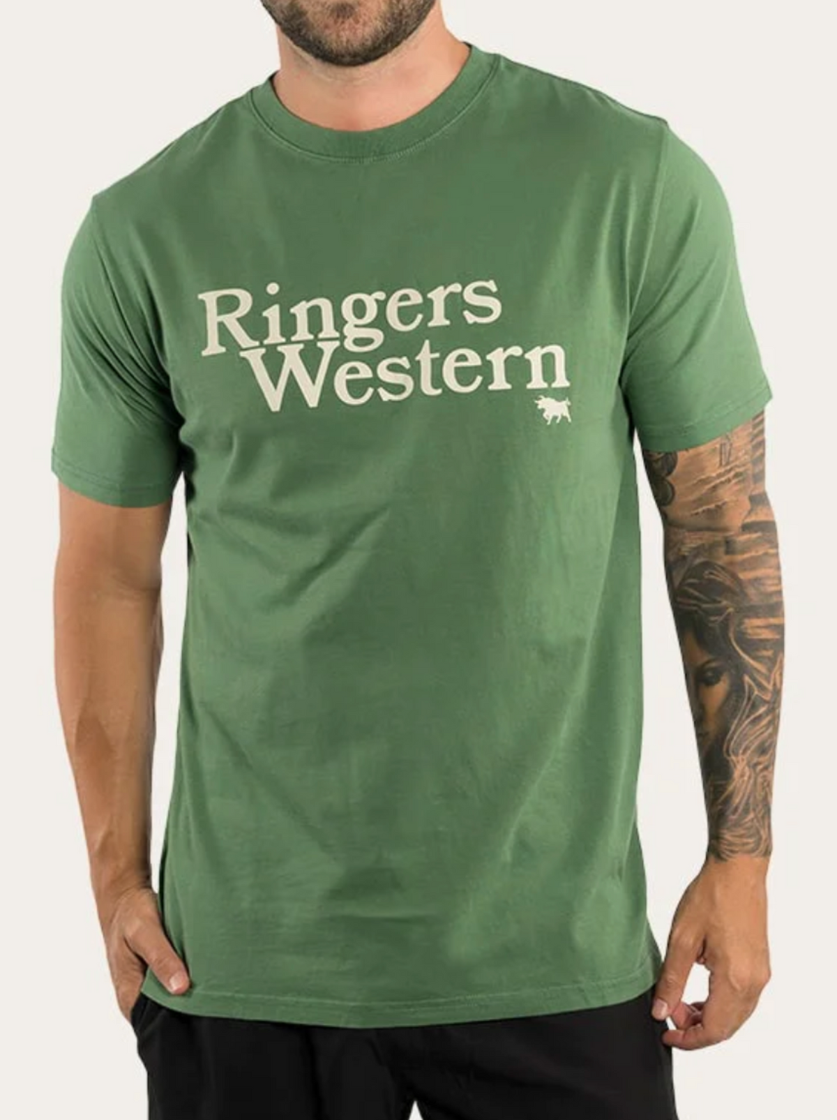 A person wearing a green Ringers Western The Lodge Classic Fit T Shirt, made from 100% cotton jersey with white text across the chest and a small bull logo underneath, stands with one hand in their pocket.