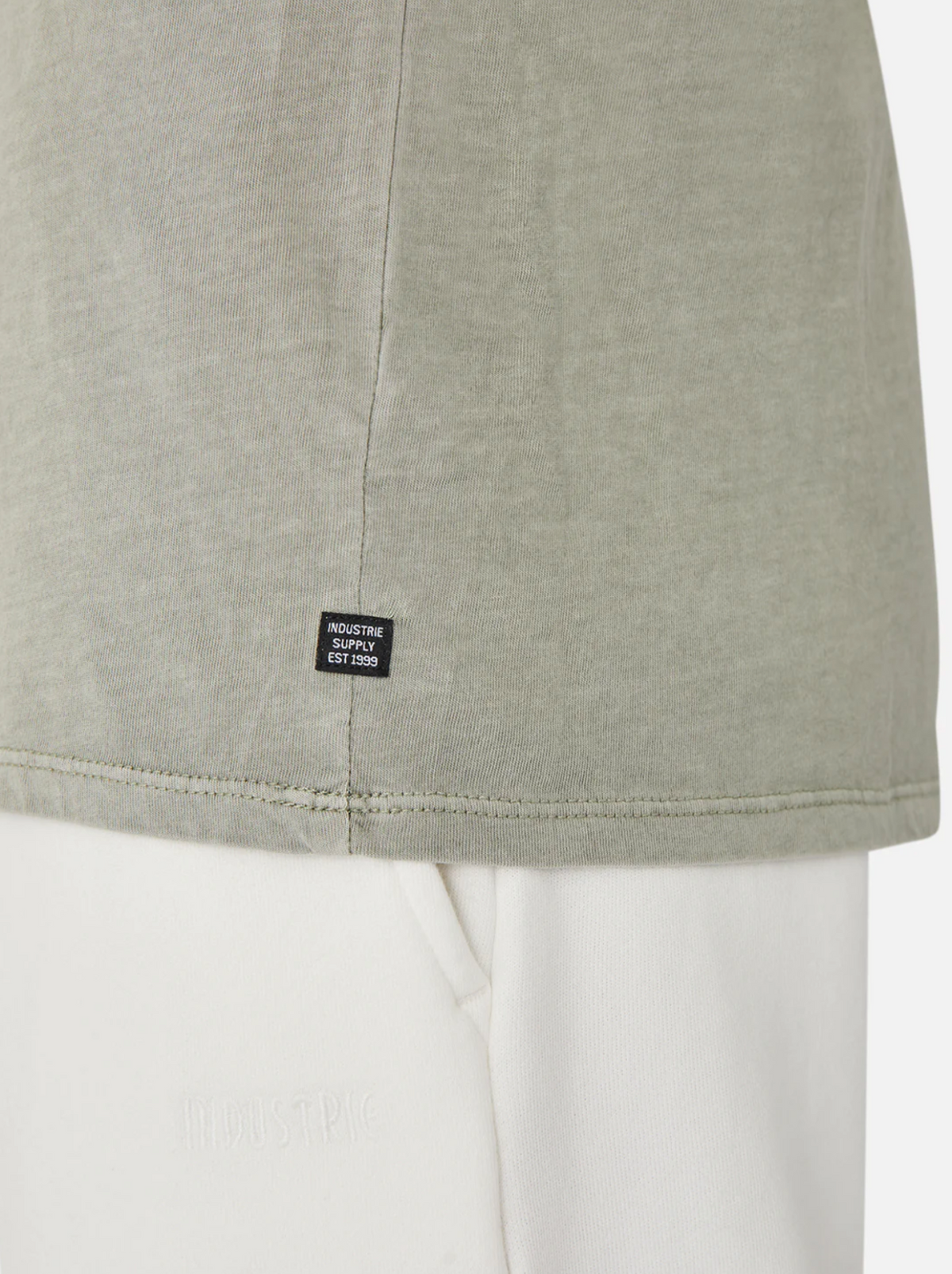 A close-up shows someone in a light green Industrie The Basic Classic Tee with a timeless fit. A small black label near the hem says, "Industrie, Established 1999." It pairs well with white pants featuring subtle text against a plain white background.