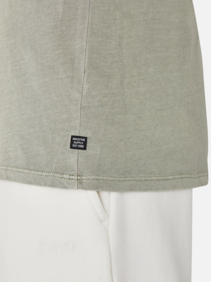 A close-up shows someone in a light green Industrie The Basic Classic Tee with a timeless fit. A small black label near the hem says, "Industrie, Established 1999." It pairs well with white pants featuring subtle text against a plain white background.