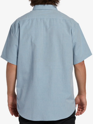 A person is shown from the back wearing a light blue Billabong All Day Short Sleeve Shirt made from woven fabric with a simple collar and dark pants; the shirt has no visible patterns or logos, embracing classic style.