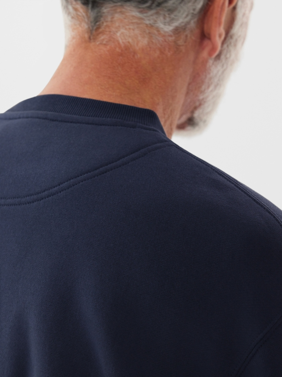 Close-up of an older man's back and shoulder wearing the RM Williams R.M.W Script Crew Neck, a cotton blend dark blue sweatshirt with an embroidered logo. His gray hair contrasts with the clothing texture and color against a plain white background.