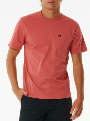 Against a white background, a person stands wearing the Rip Curl Plain Pocket Tee in classic staple coral, paired with black shorts. The short-sleeve shirt features a left chest pocket with a small black tag.