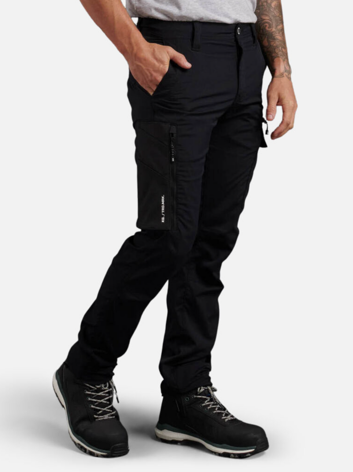 A person wearing black King Gee Trademark Cargo Pants, a grey t-shirt, and black work boots stands with one hand in their pocket; a tattoo is visible on their left forearm.