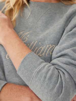 A close-up shows a person in a gray RM Williams R.M.W Womens Script Crew sweatshirt. The cursive script is partially visible, with their bent arm highlighting the sleeve texture. Blonde hair can be seen at the top of the image, enhancing their functional style.