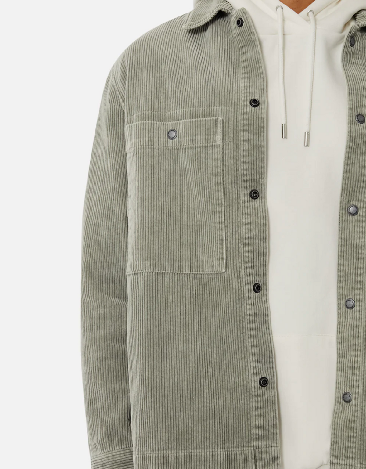 A person wears the Industrie The Atmore Jacket—a light grey-green 100% cotton corduroy button-up with chest pocket and black buttons—layered over a white hoodie. Face is not visible.