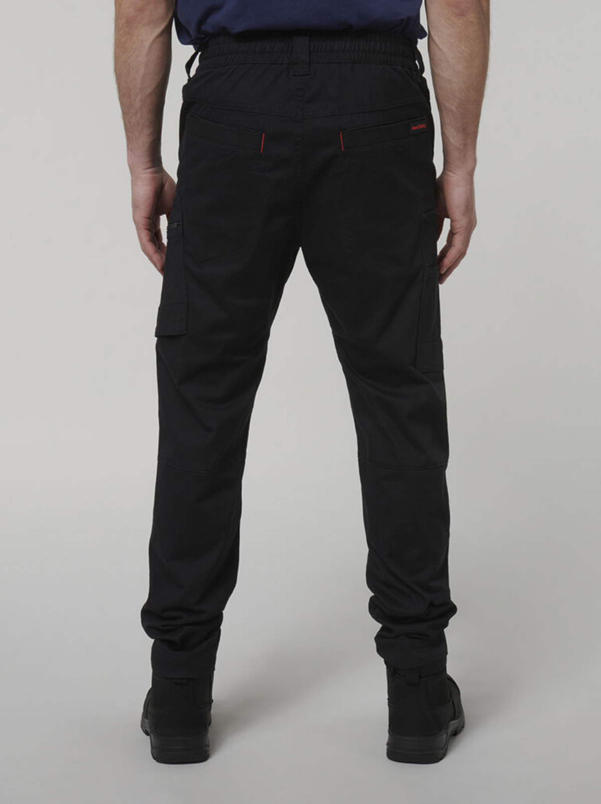 A person stands with their back to the camera, wearing black Hard Yakka Tough Maxx Pant cargo pants and black shoes against a plain light gray background.