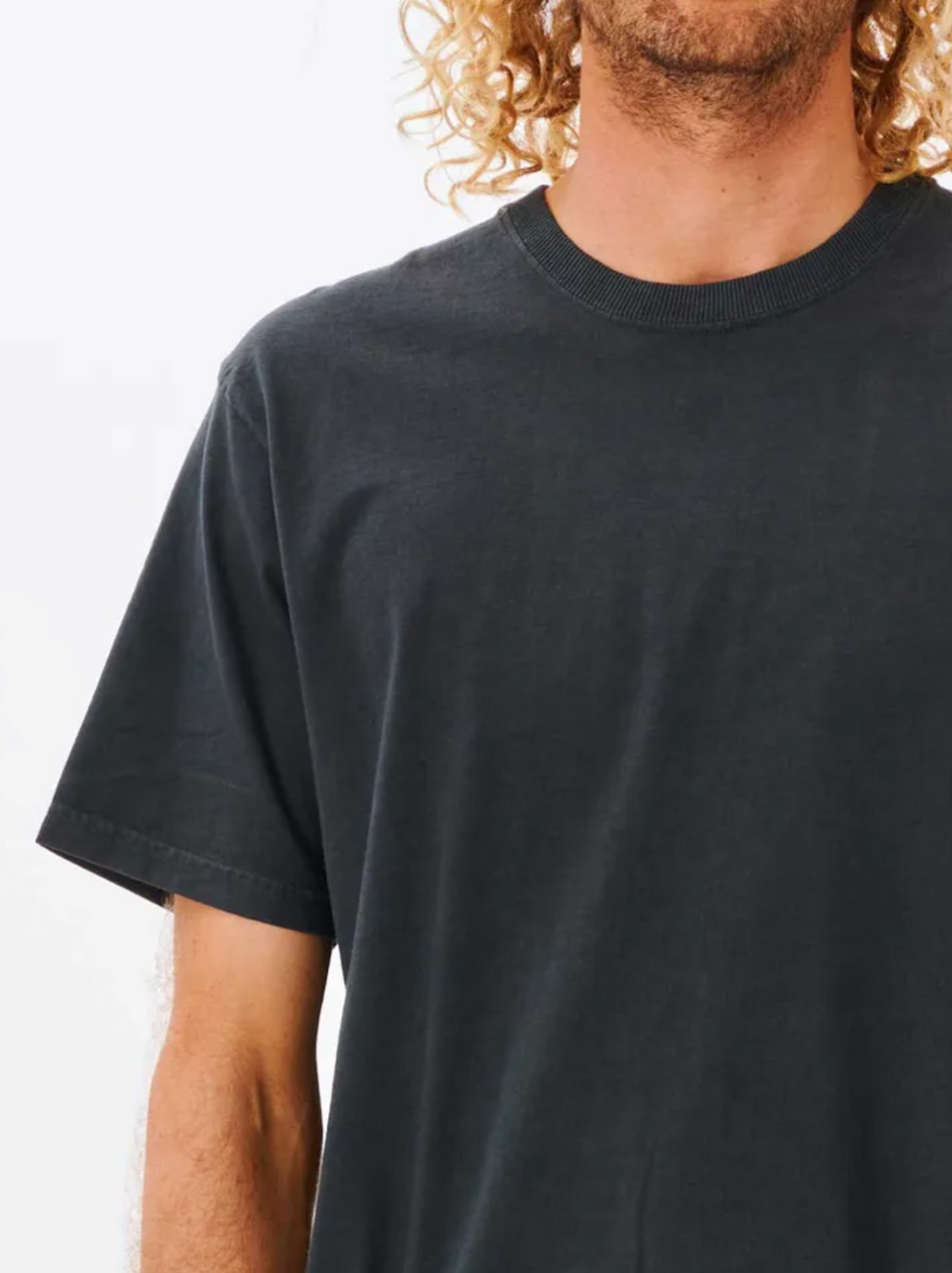 A person with curly blond hair wears a black Rip Curl Plain Wash Tee, relaxed fit and pigment dyed, shown from the waist up against a white background.