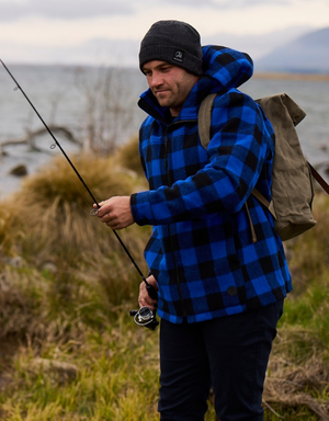 A person in a blue and black checkered Swanndri Hudson Hoody and grey beanie is standing near a body of water, holding a fishing rod. They have a brown backpack and are surrounded by grass and rocks with mountains in the background.