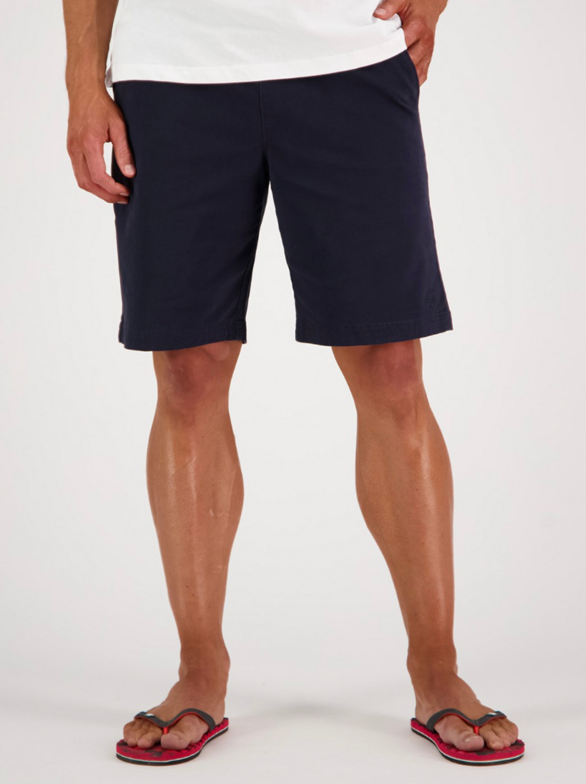 A person in a white t-shirt, Swanndri Clevedon Stretch Canvas Shorts, and pink flip-flops stands against a light background with relaxed hands by their sides.