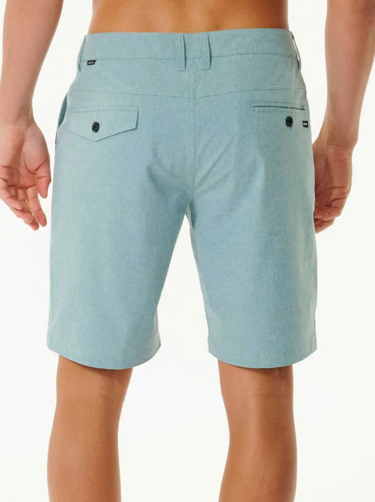 A person is seen from behind wearing light blue Rip Curl Boardwalk Phase Nineteen Shorts with belt loops, a left buttoned flap back pocket, and a right welt pocket with button, set against a plain light background.