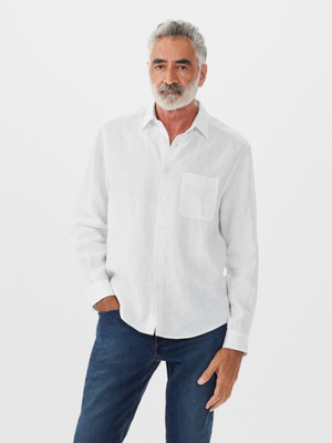 A grey-haired man with a beard wears a white RM Williams Coalcliff Shirt and blue jeans, one hand in his pocket, confidently posing against a plain white background.