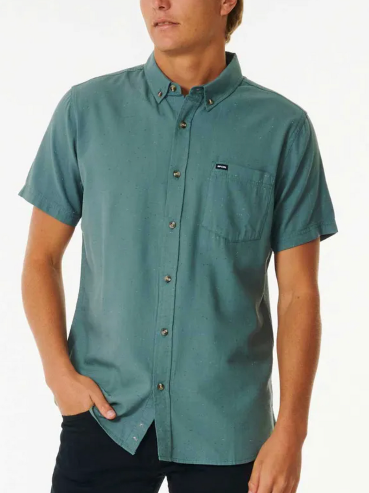 A person dons a Rip Curl Ourtime Short Sleeve Shirt, a teal button-up featuring subtle dots and a chest pocket, perfectly matched with black pants against a plain white background.