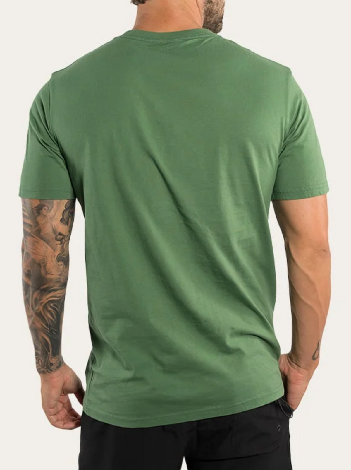 A man stands with his back to the camera, wearing a Ringers Western The Lodge Classic Fit T Shirt in plain green, made from 100% cotton jersey, and black pants. His right arm features tattoos and his hands rest relaxed by his sides.
