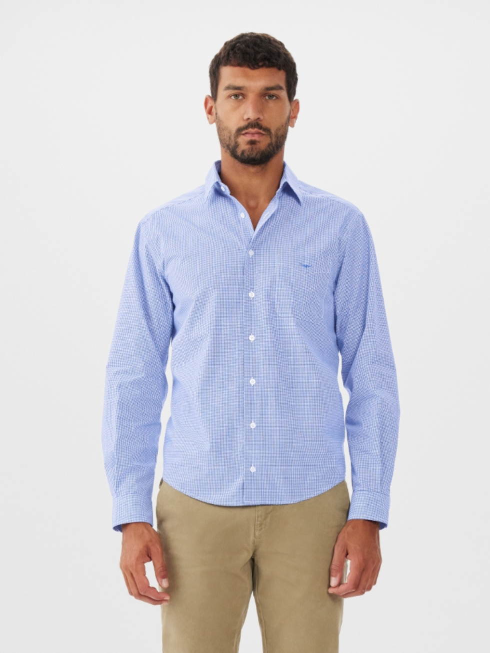 A man with short dark hair and a beard wears the RM Williams Coalcliff Shirt in light blue check with subtle longhorn embroidery and khaki pants, standing against a plain white background.