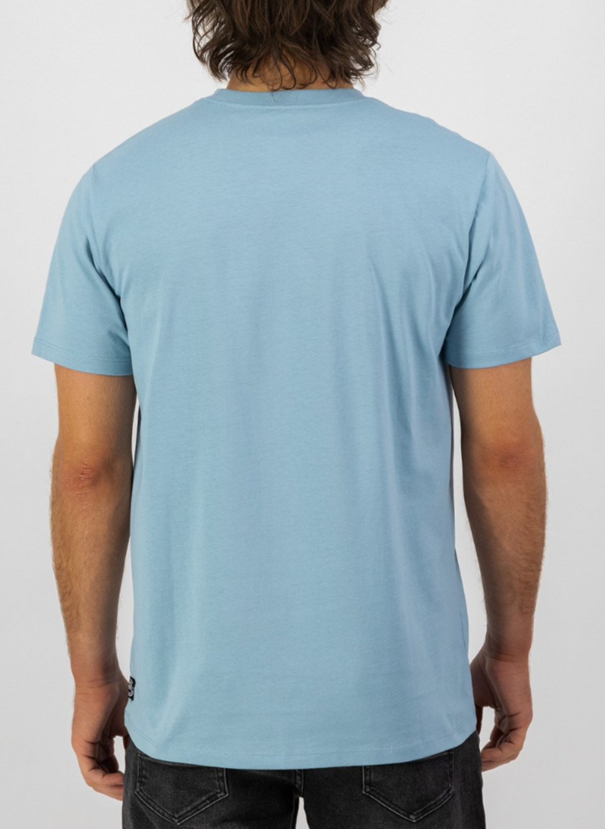 A person with medium-length brown hair stands facing away from the camera, wearing a Swanndri Original v2 Print T-Shirt in light blue and black jeans, set against a plain white background.