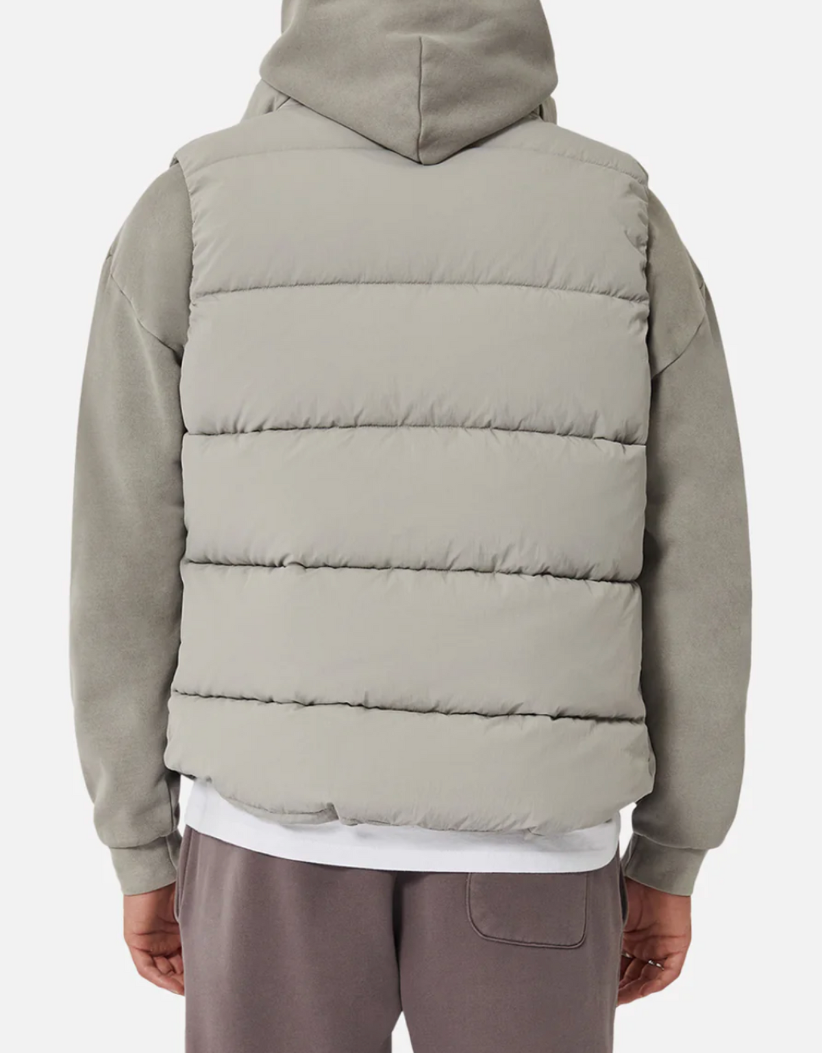 A person is seen from behind, wearing the Industrie The New Hanam Puffer Vest in light gray, which sports thick quilted padding and horizontal stitching over a gray hoodie with the hood up. The vest is crafted from durable Nylon/Polyester material. Additionally, the person is wearing brownish-gray pants against a plain white background.