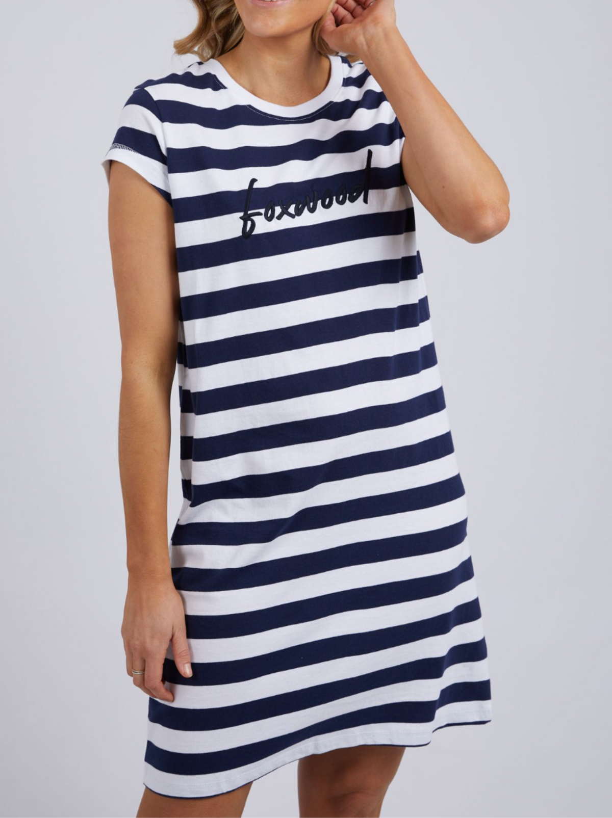The woman is wearing a Foxwood Signature Stripe Tee Dress, crafted from 100% cotton jersey with navy and white stripes. The word "bosswood" is prominently displayed on the front as she poses against a plain backdrop.