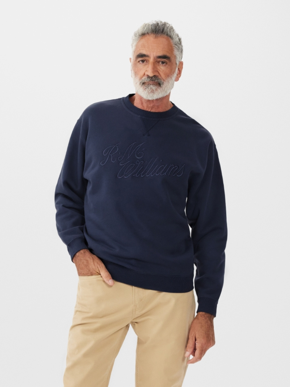 An older man with a gray beard and hair stands against a white background, wearing the RM Williams R.M.W Script Crew Neck in navy blue and tan pants, with his left hand in his pocket.