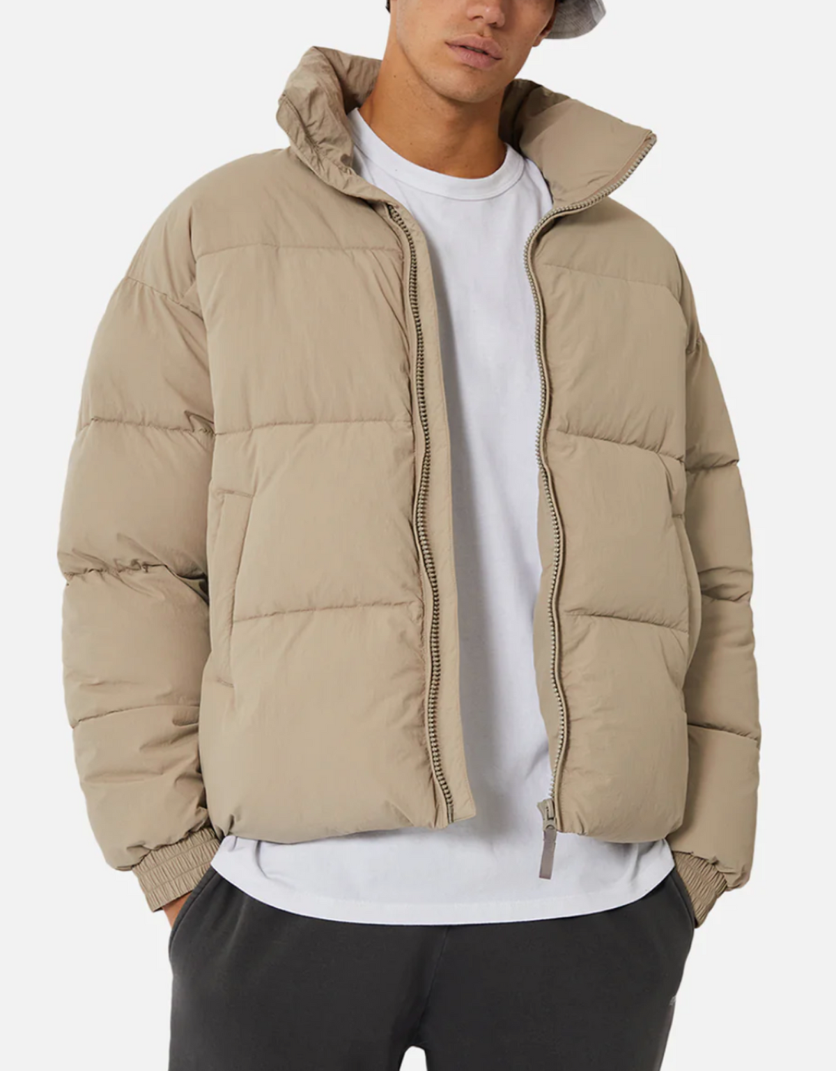 A person wearing Industrie The Regent Puffer Jacket in beige with quilted padding over a white t-shirt and black pants. They are also sporting a light grey bucket hat. The jacket, featuring an Industrie label, is unzipped, and the person's hands rest in their pant pockets against a plain white background.