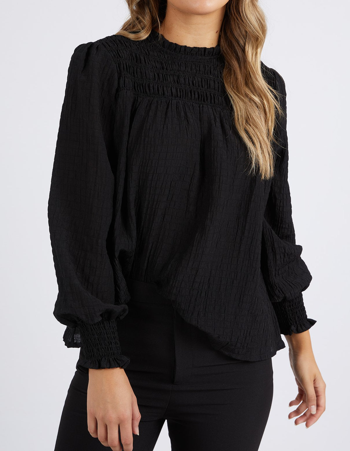 A woman wears the Foxwood Helena Check Top—a long-sleeve, textured black blouse with a gathered neckline and cuffs—paired with black pants. She has wavy light brown hair and stands against a plain white background.