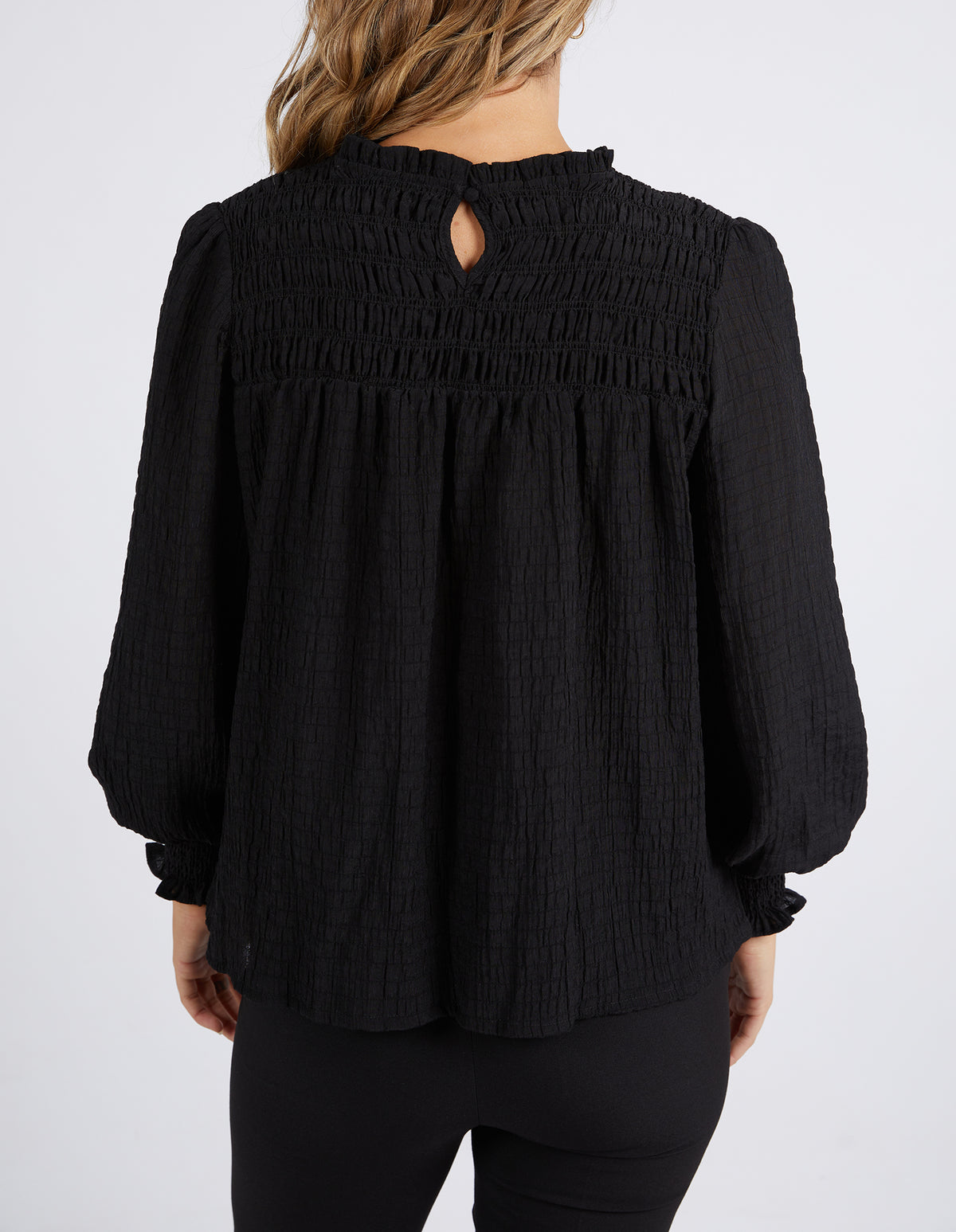 A woman with wavy, light brown hair is seen from the back wearing the Foxwood Helena Check Top, which has a relaxed fit, gathered yoke, and button keyhole at the neckline. She pairs it with black pants.