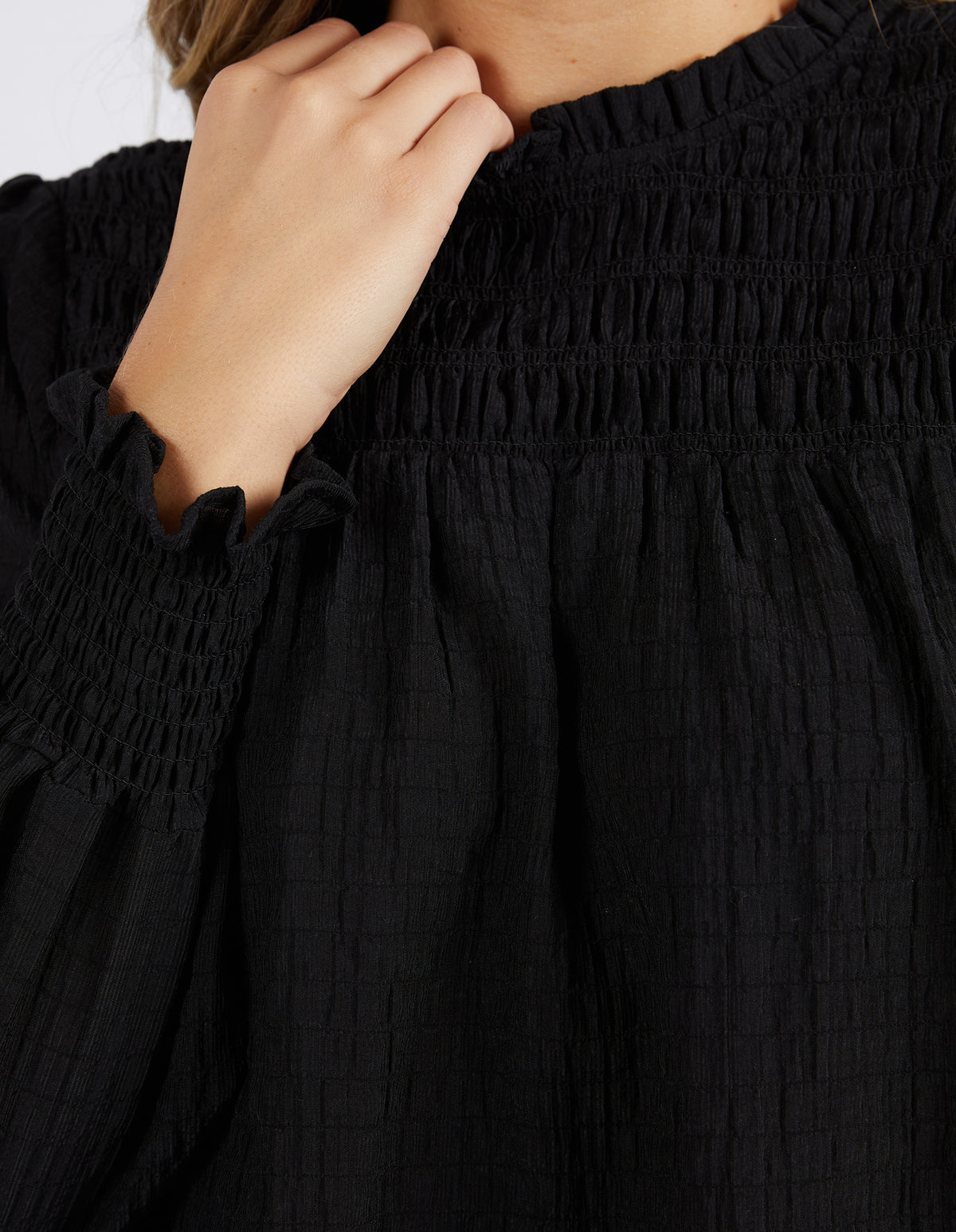 Wearing the Foxwood Helena Check Top—a black, long-sleeve blouse with a gathered neckline and ruffled cuffs. The relaxed-fit, smocked top is styled as their hand touches their face, only the lower half visible.