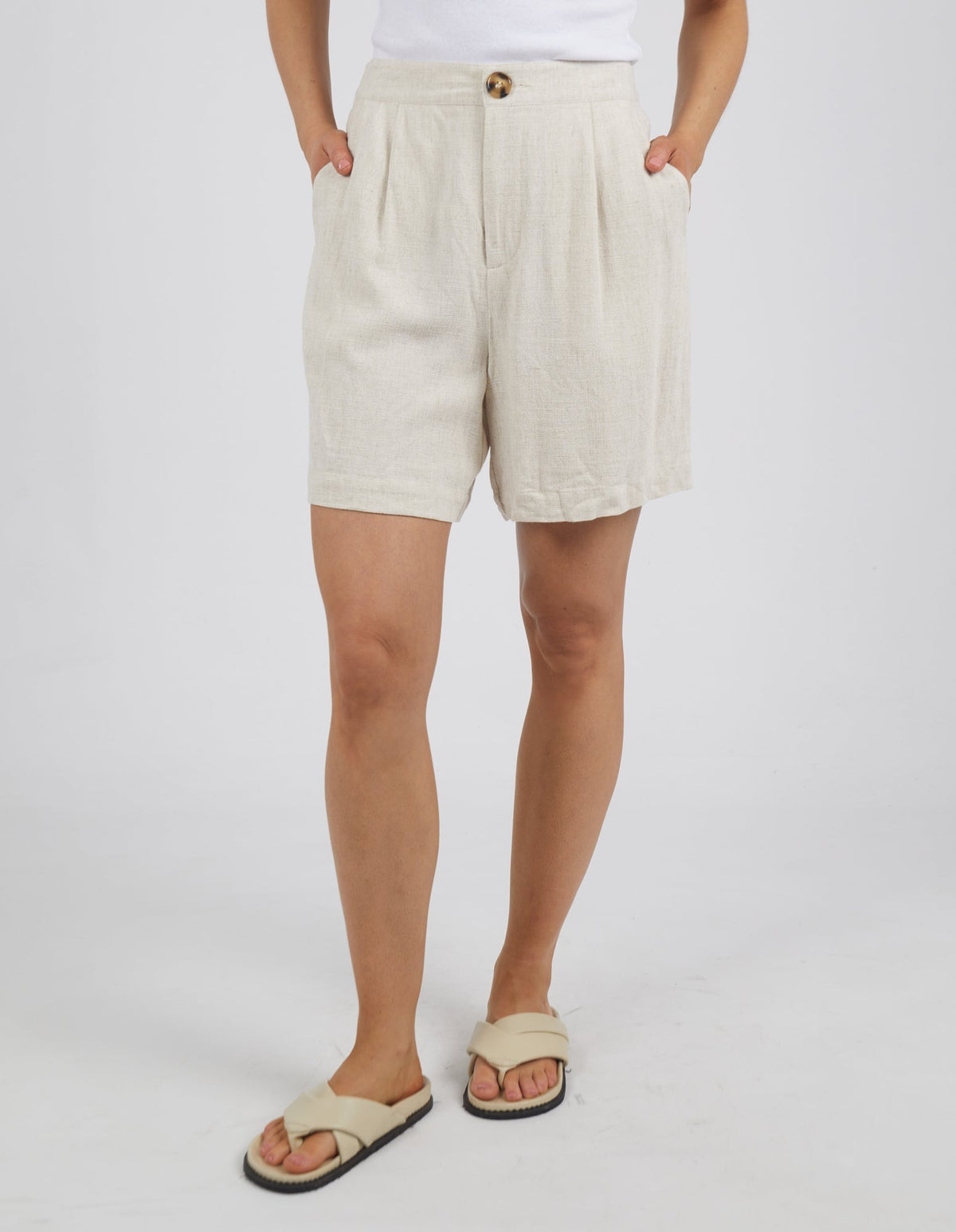 In Foxwood Farrow Short with beige pleats and a brown button, a person pairs it with a white top and beige sandals. With hands in pockets, they stand against a plain white background, highlighting the mid-thigh length elegance.
