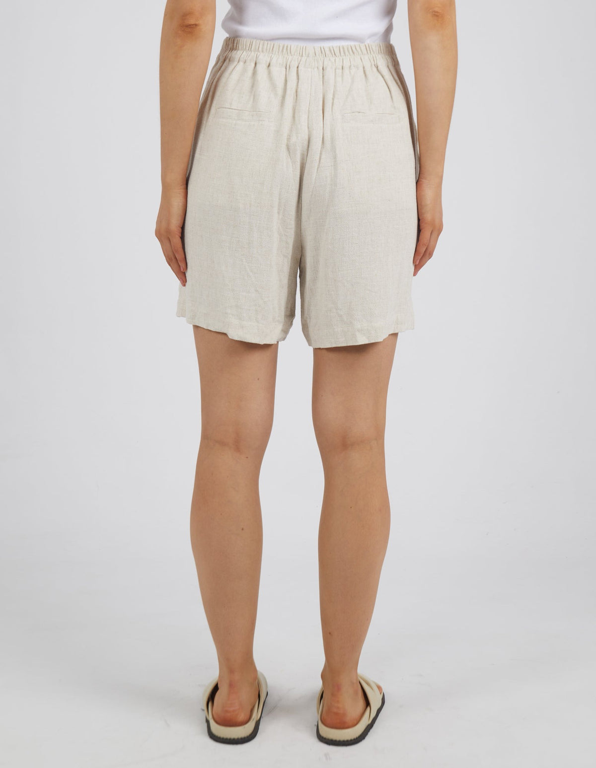 A person is seen from behind wearing Foxwood Farrow Shorts featuring an elastic waistband and two pockets. They are matched with a white shirt over a neutral backdrop, accessorized with sandals for a comfy fit.