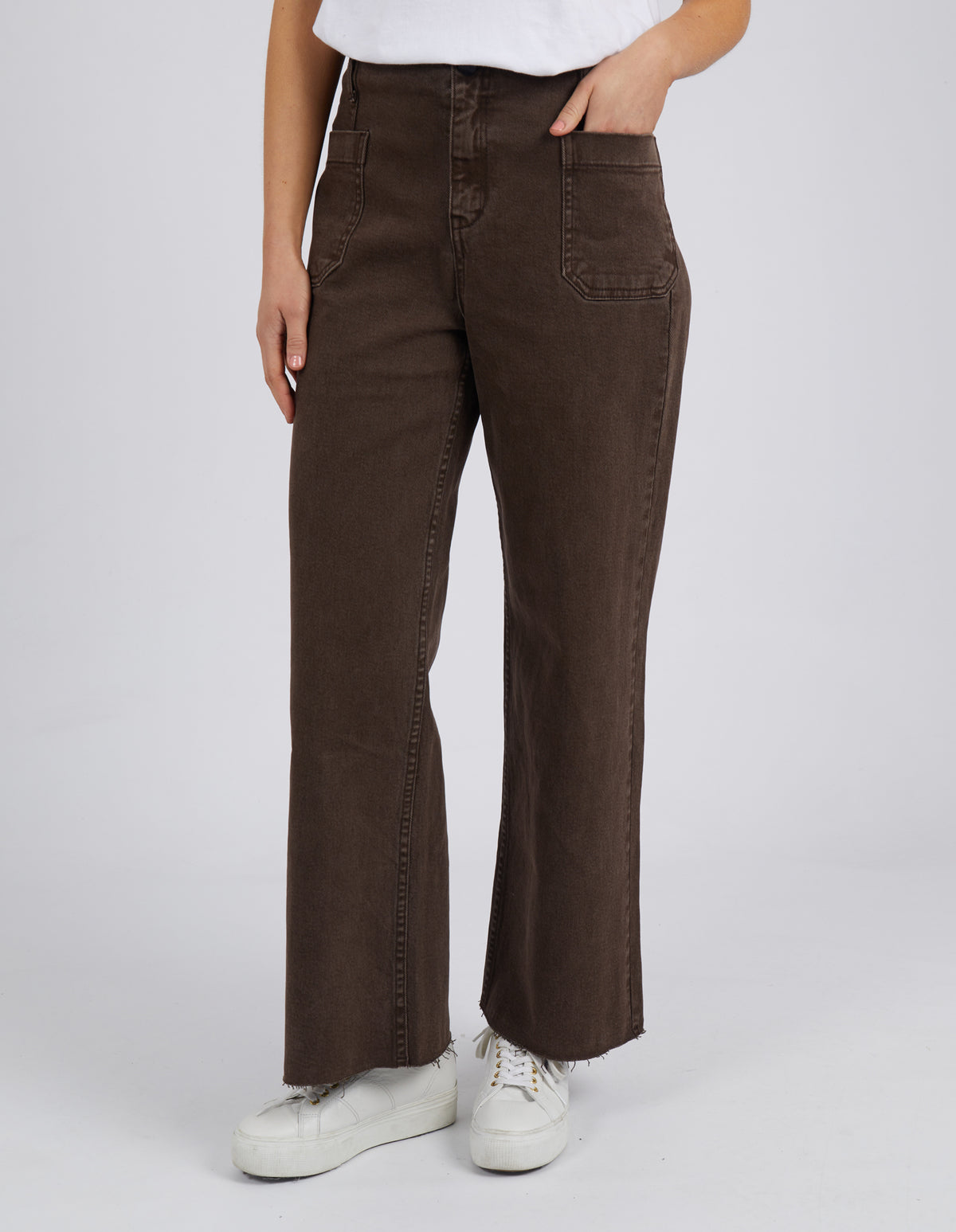 A person stands confidently in the Foxwood Eddie Wide Leg Jean, featuring large front pockets and raw edge detailing, paired with a crisp white shirt and sneakers. The plain light gray background complements this effortlessly stylish look.