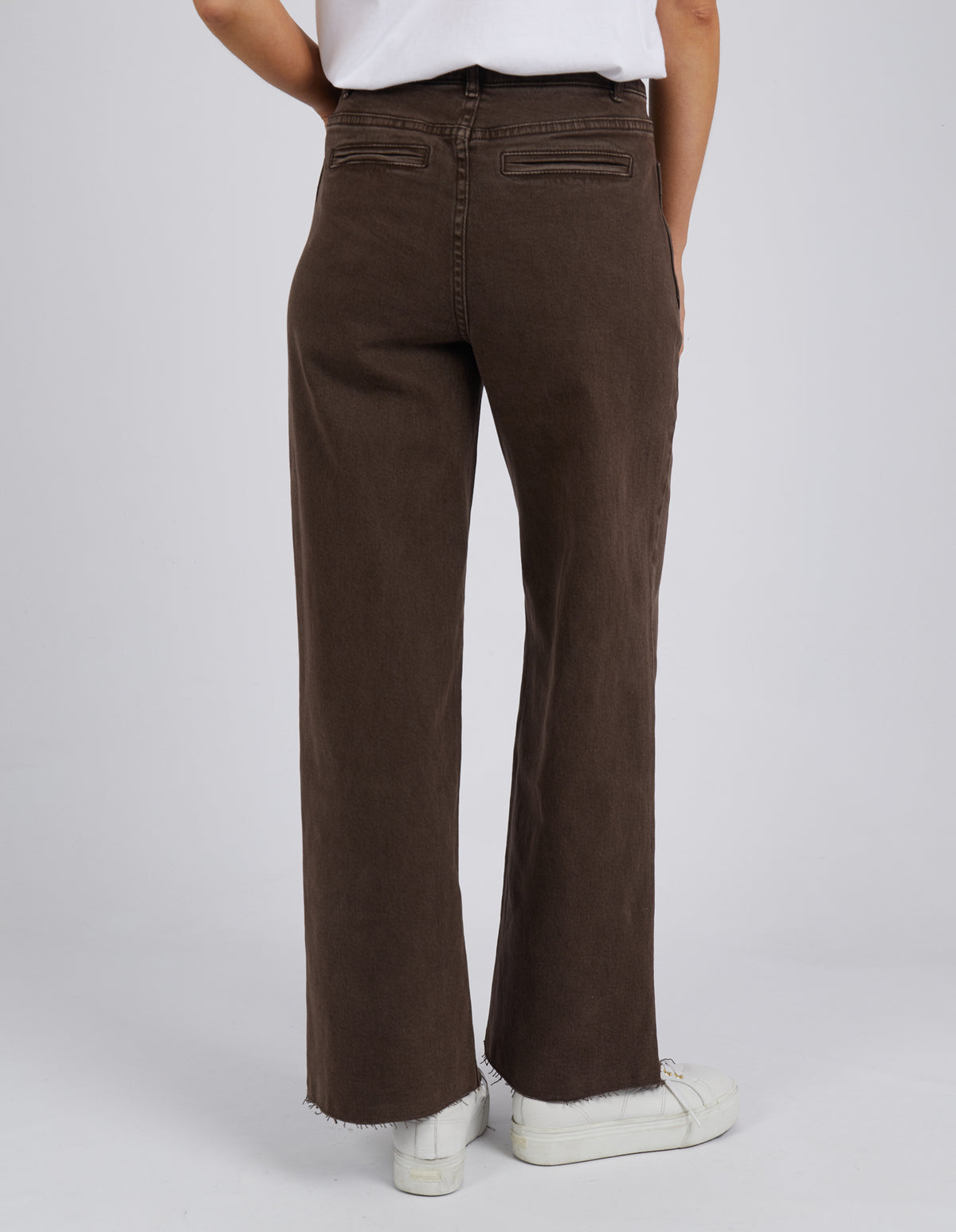 A person wearing Foxwood Eddie Wide Leg Jeans in dark brown, featuring raw edge detailing, pairs them stylishly with a white top and comfortable white sneakers while standing on a light grey floor.