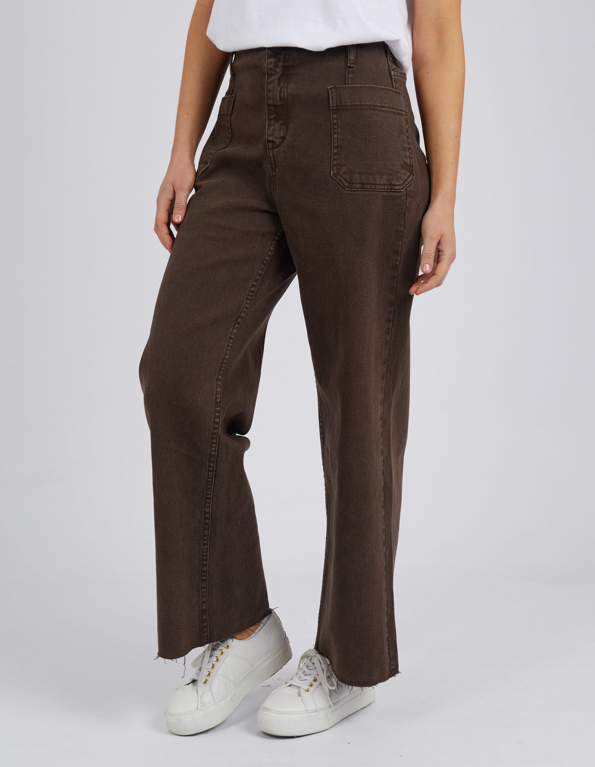 A person wears Foxwood Eddie Wide Leg Jeans, featuring a high waist, wide legs, and raw edge detailing. Paired with a white t-shirt and white sneakers, the jeans with front pockets create a stylish ensemble against a plain gray background.