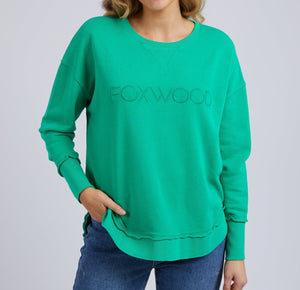 A woman models the Foxwood Simplified Crew, a bright green 100% cotton sweatshirt with "FOXWOOD" embroidery on the front, styled with blue jeans. She stands against a light gray background, her hair loose and one hand in her pocket.