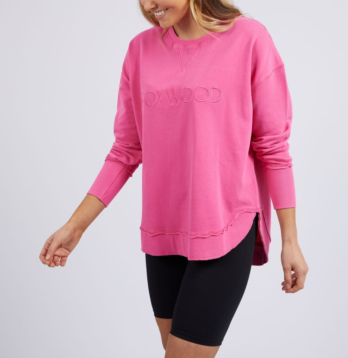 A woman wears the bright pink Foxwood Simplified Crew, featuring "FOXWOOD" embroidery on the front, and black bike shorts while smiling and posing against a plain light background.